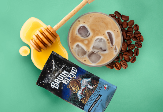 An icy, frothy glass of creamy shaken coffee surrounded by coffee beans, honey and a package of Bruin Blend Coffee.