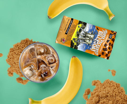 An icy glass of creamy Shaken Iced Banana Bread Espresso sitting atop piles of brown sugar and surrounded by bananas and a package of Three Peckered Billy Goat Coffee.