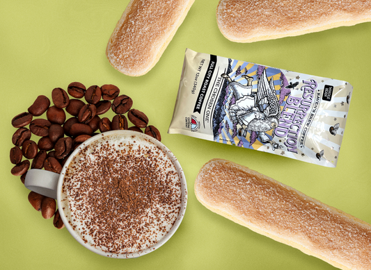 Image of a mug of creamy tiramisu latte sprinkled with coco powder atop a pile of beans. Larger than life lady fingers and a small Resurrection Blend package surround the coffee mug.