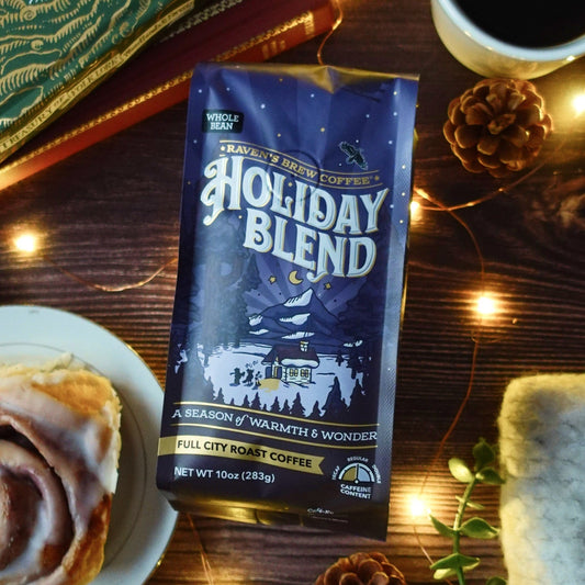 Raven's Brew Coffee 10oz Holiday Blend French Roast Whole Bean Coffee package on a wooden table with a cinnamon roll and coffee cup.