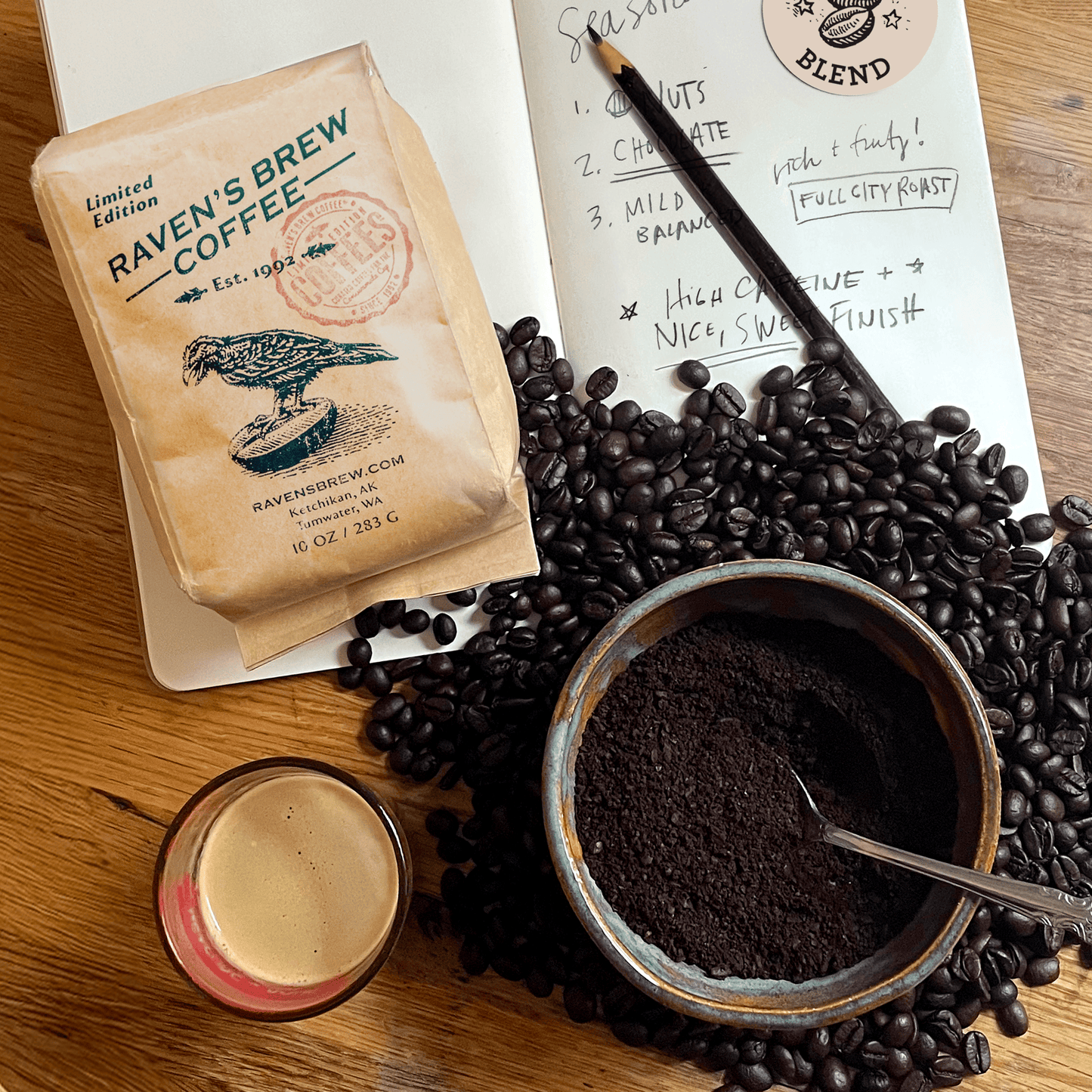 Kraft bag of Roaster's Pick: Seasonal Blend on a table with open sketch book, a pile of beans, a bowl with coffee grounds, and a freshly pulled shot laid on top.