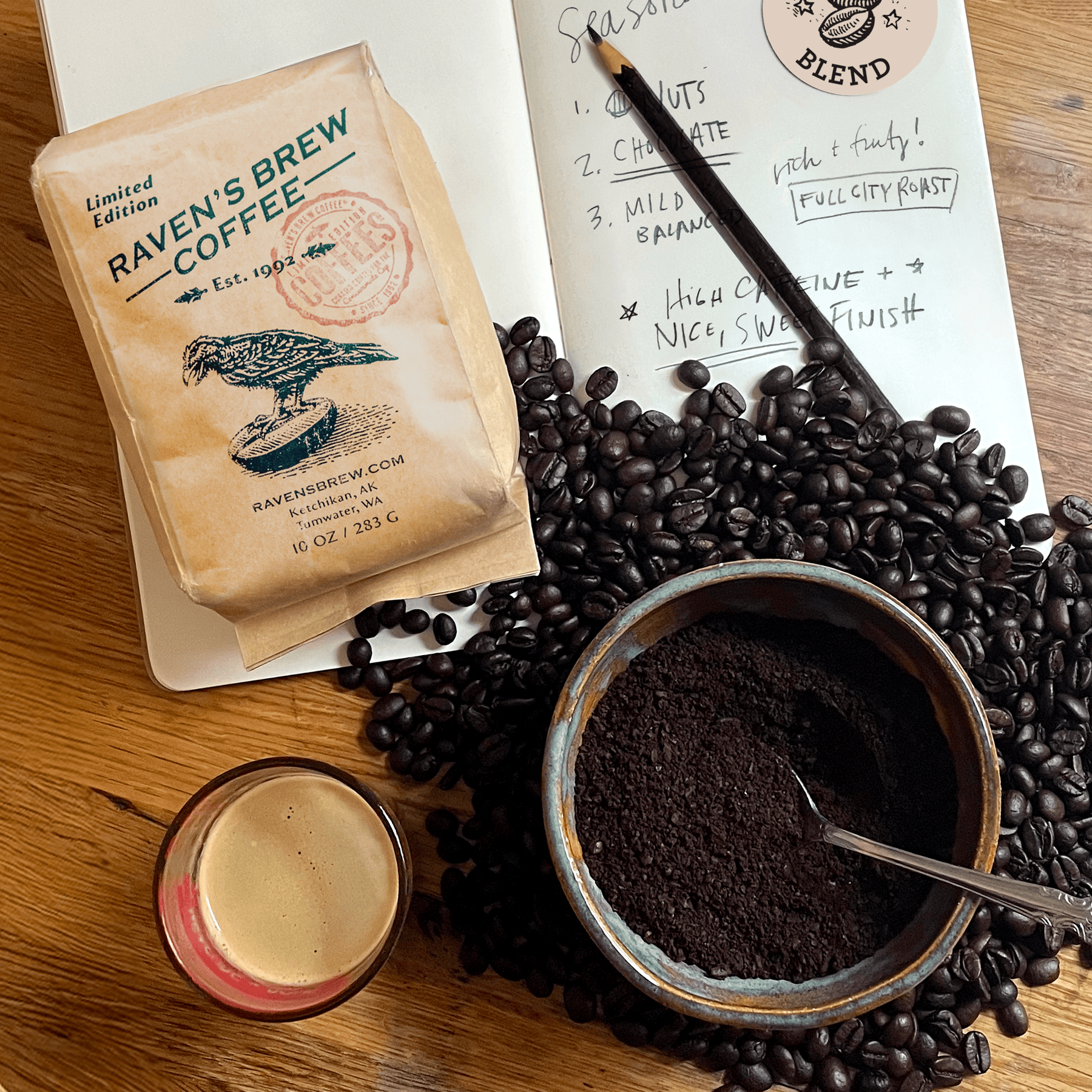 Kraft bag of Roaster's Pick: Seasonal Blend on a table with open sketch book, a pile of beans, a bowl with coffee grounds, and a freshly pulled shot laid on top.