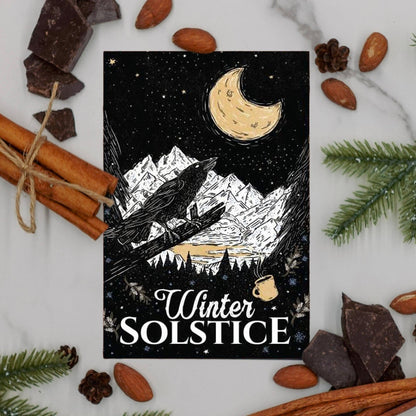 Winter Solstice Coffee postcard  surrounded by cinnamon sticks, chocolate pieces, and pine cones on a marble surface.