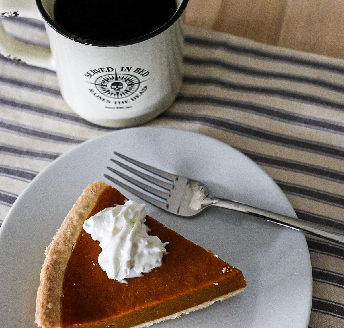 Slice of pumpkin pie with whipped cream on a plate with a fork, next to a mug on a striped cloth.